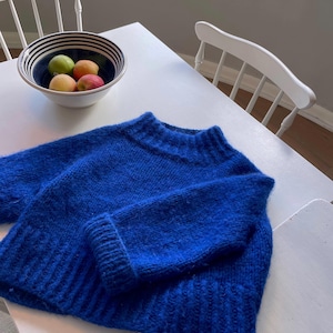 Pode incluir: Um suéter azul brilhante e felpudo com gola alta e mangas compridas. O suéter está dobrado e colocado sobre uma mesa branca.