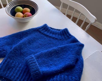 Puede incluir: Un suéter azul brillante y peludo con cuello alto y mangas largas. El suéter está doblado y colocado sobre una mesa blanca.
