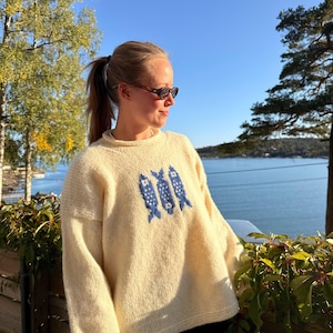 K&ouml;nnte beinhalten: Ein cremefarbener, lang&auml;rmeliger Strickpullover mit Rundhalsausschnitt und einem zentralen Grafikdesign aus drei stilisierten blauen Fischen. Der Pullover hat eine lockere Passform mit gerippten B&uuml;ndchen und Saum aus weichem Garn.