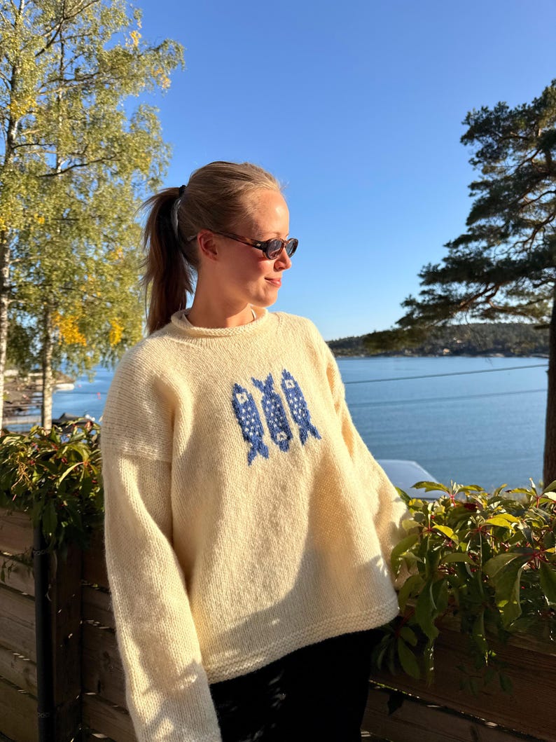 K&ouml;nnte beinhalten: Ein cremefarbener, lang&auml;rmeliger Strickpullover mit Rundhalsausschnitt und einem zentralen Grafikdesign aus drei stilisierten blauen Fischen. Der Pullover hat eine lockere Passform mit gerippten B&uuml;ndchen und Saum aus weichem Garn.