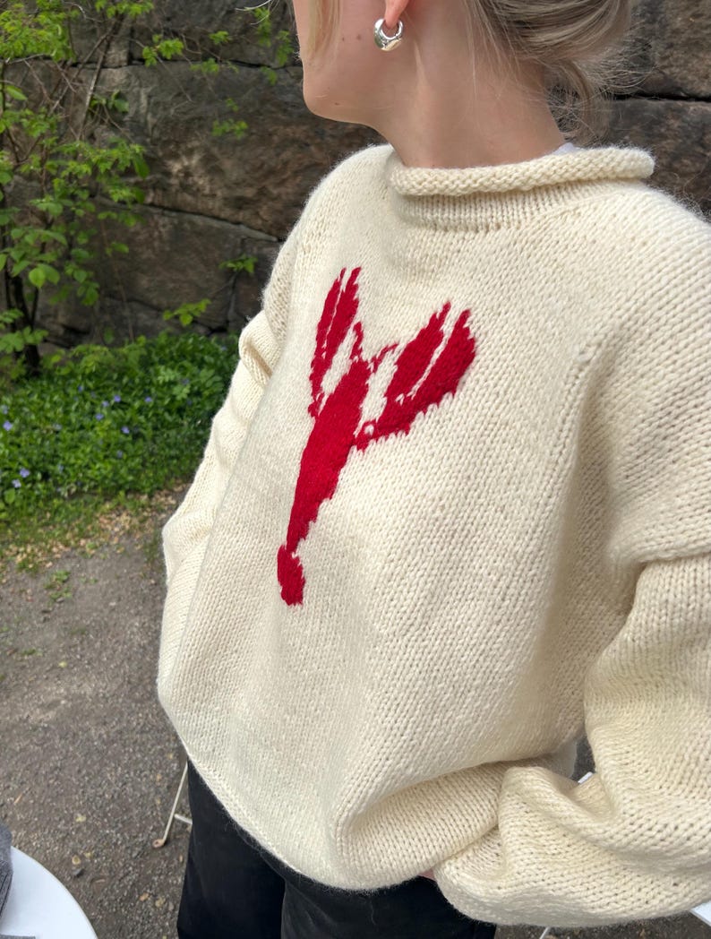 K&ouml;nnte beinhalten: Ein cremefarbener Langarm-Strickpullover mit einer markanten roten Hummer-Grafik in der Mitte der Brust. Der Pullover hat einen dicken, gerippten Rundhalsausschnitt und eine grobe Strickstruktur, die f&uuml;r eine lockere Passform sorgt.