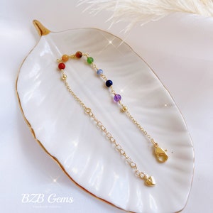 May include: A gold chain bracelet with a heart charm and seven colorful gemstone beads. The beads are red, orange, yellow, green, blue, indigo, and violet. The bracelet is on a white plate with a gold rim.