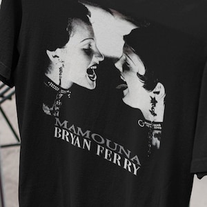 Könnte beinhalten: Schwarzes T-Shirt mit einer Schwarz-Weiß-Grafik von zwei Frauen, die sich gegenüberstehen und den Mund geöffnet haben. Der Text "MAMOUNA BRYAN FERRY" ist darunter gedruckt. Das T-Shirt hängt an einem Holzbügel.