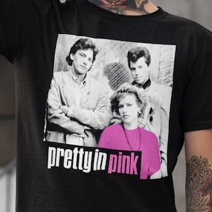 May include: Black t-shirt with a graphic from the film "Pretty in Pink." The graphic shows the cast in black and white, with the words "pretty in pink" in white. The shirt is worn by a person with tattoos and green hair.