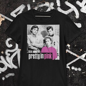 May include: Black t-shirt with a graphic from the film "Pretty in Pink." The graphic shows three people in a black and white photo with the movie title in pink and white text. The shirt is hanging on a gold hanger.