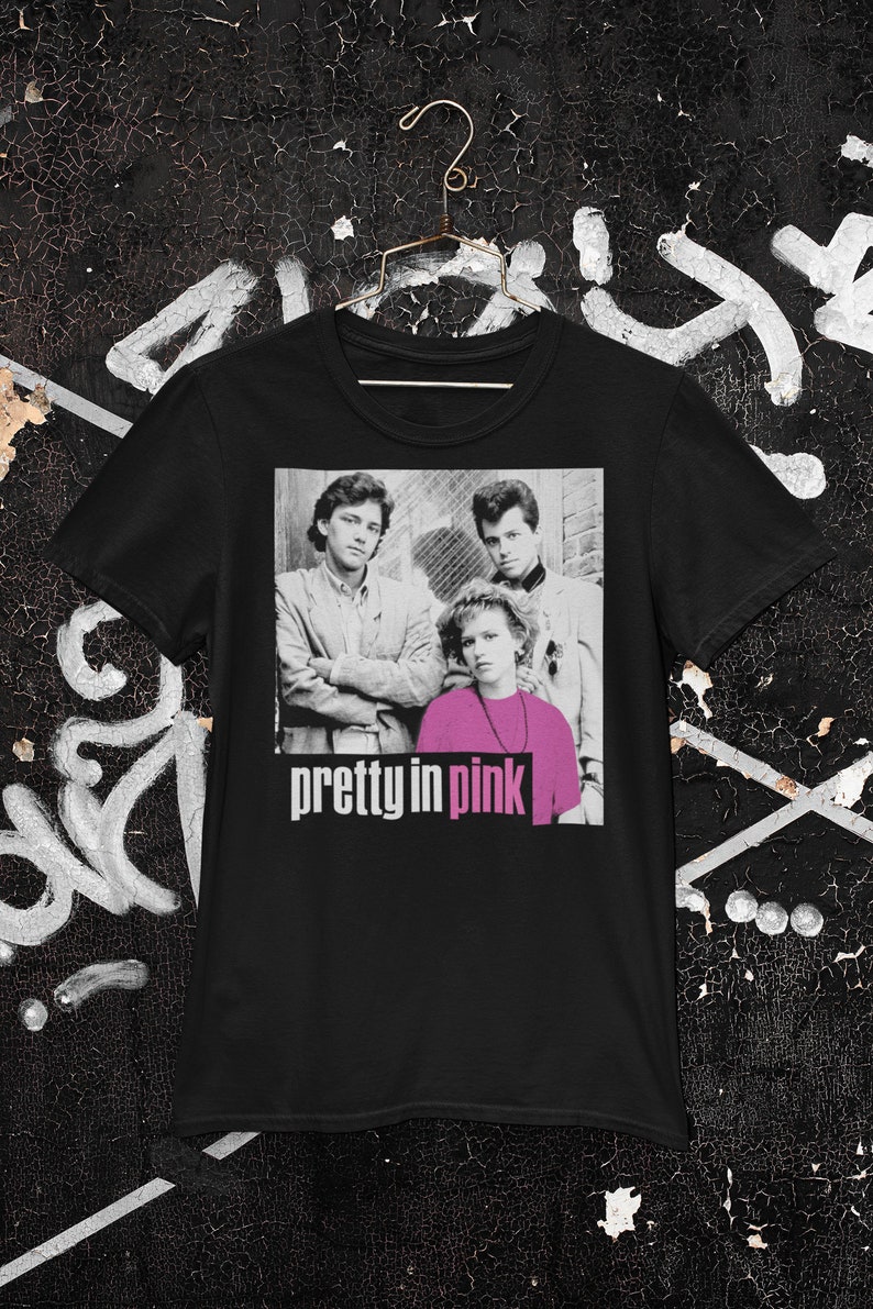 May include: Black t-shirt with a graphic from the film "Pretty in Pink." The graphic shows three people in a black and white photo with the movie title in pink and white text. The shirt is hanging on a gold hanger.