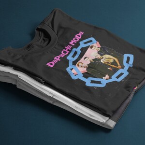 May include: Black t-shirt featuring a band photo and the text "DEPECHE MODE" in pink and blue. The design includes a chain-link border around the band members. The shirt is folded on top of other folded shirts.