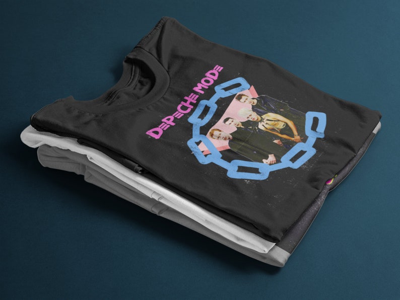 May include: Black t-shirt featuring a band photo and the text "DEPECHE MODE" in pink and blue. The design includes a chain-link border around the band members. The shirt is folded on top of other folded shirts.
