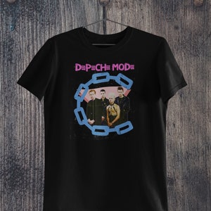 May include: Black t-shirt featuring the band Depeche Mode. The shirt has the band's name in pink and a blue chain design around a photo of the band members. The shirt is hanging on a wooden background.