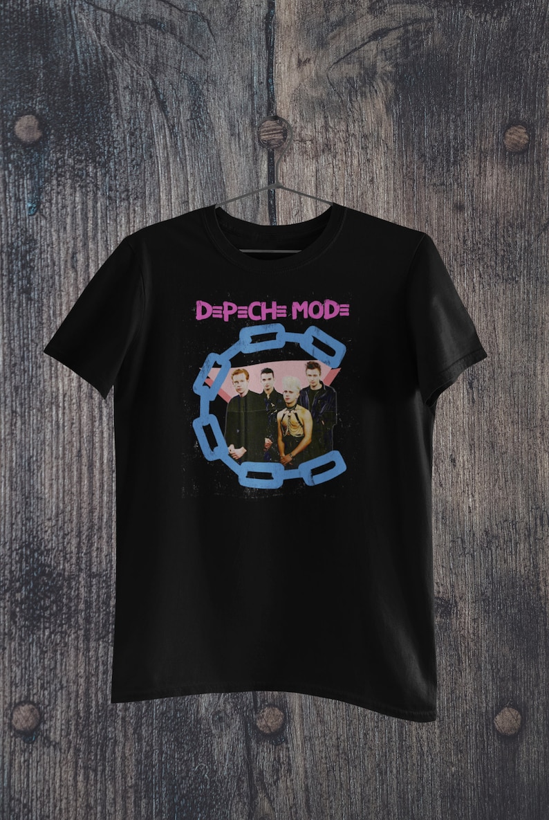 May include: Black t-shirt featuring the band Depeche Mode. The shirt has the band's name in pink and a blue chain design around a photo of the band members. The shirt is hanging on a wooden background.