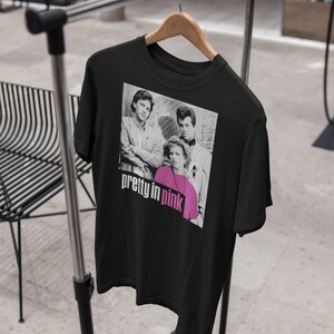 May include: Black t-shirt with a black and white image of three people and the text "pretty in pink" in pink and white. The t-shirt is displayed on a wooden hanger on a metal clothing rack.