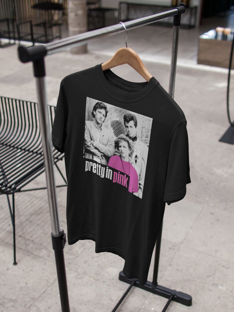 May include: Black t-shirt with a black and white image of three people and the text "pretty in pink" in pink and white. The t-shirt is displayed on a wooden hanger on a metal clothing rack.