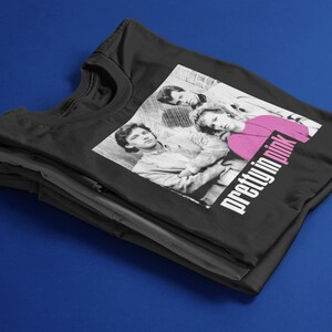May include: Black t-shirt with a black and white image of three people and the text "pretty in pink" in white and pink. The t-shirt is folded on top of other black and grey t-shirts against a blue background.
