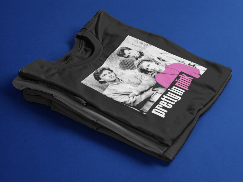May include: Black t-shirt with a black and white image of three people and the text "pretty in pink" in white and pink. The t-shirt is folded on top of other black and grey t-shirts against a blue background.