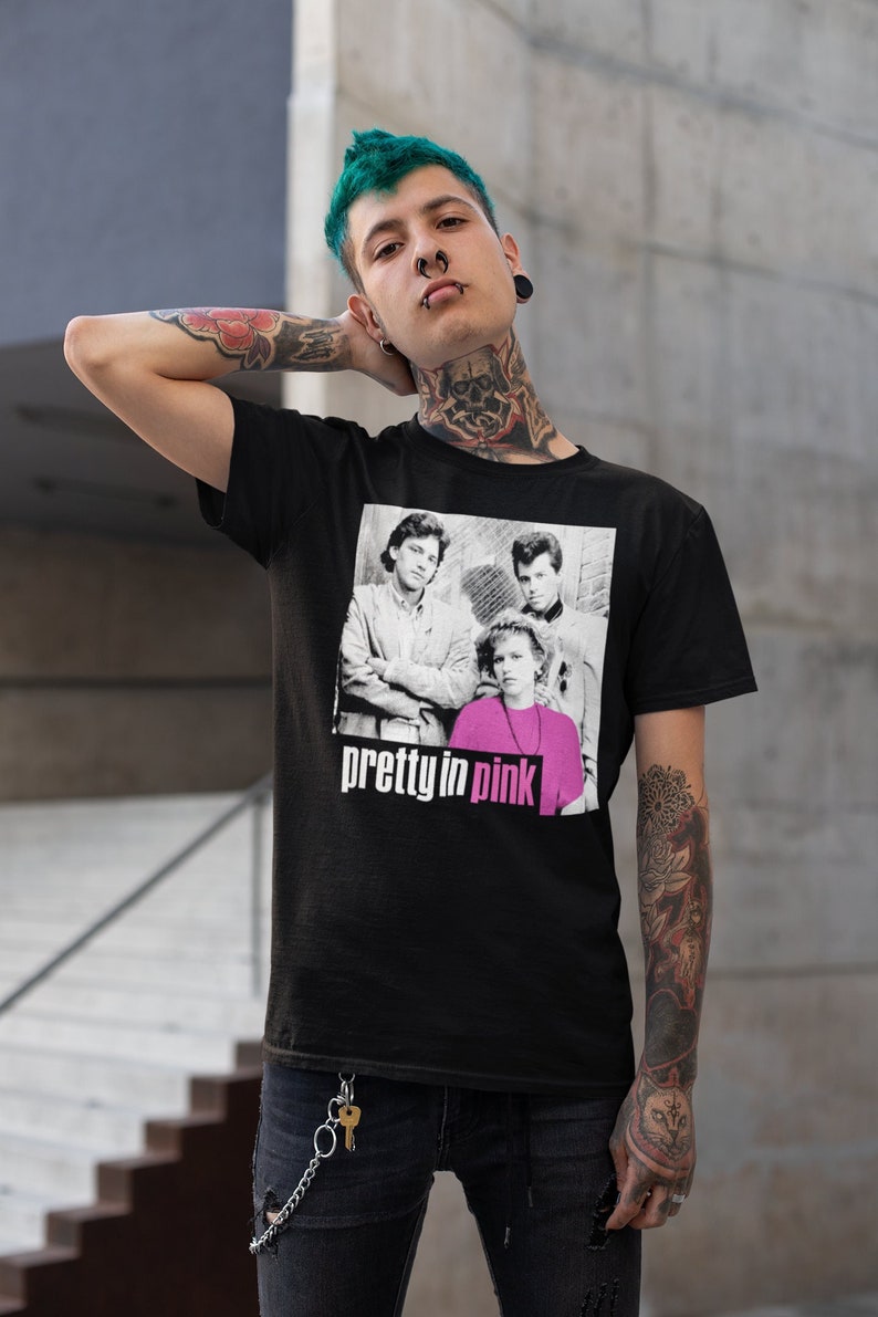 May include: Black t-shirt with a graphic from the film "Pretty in Pink." The graphic shows the cast in black and white, with the words "pretty in pink" in white. The shirt is worn by a person with tattoos and green hair.