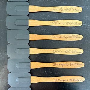 May include: Eight wooden handled silicone spatulas with personalised text. The text reads "Mum's Kitchen", "Dad's Kitchen", "Nana's Kitchen", "Grandma's Kitchen", "Pa's Kitchen", "Grandpa's Kitchen", "Magic Maker", and "Happiness is homemade".