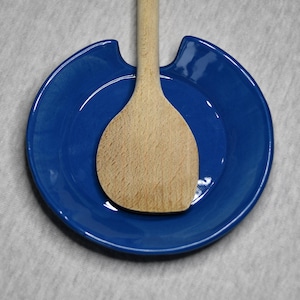 Può includere: Un poggia-cucchiaio in ceramica blu con un cucchiaio di legno al suo interno. Il poggia-cucchiaio ha una forma curva con un'apertura per il manico del cucchiaio.