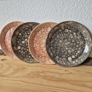 Peut inclure: Quatre assiettes en céramique avec un design texturé unique. Les assiettes présentent un motif en forme de bulles dans des tons de rose, de marron et de gris. Elles sont disposées sur une surface en bois, mettant en valeur leur attrait artistique et décoratif.