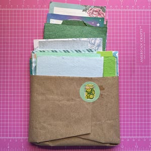 May include: A brown paper envelope filled with a variety of paper scraps in shades of green, blue, and white, featuring floral and textured patterns. A circular sticker depicting a glass jar filled with sliced cucumbers is centered on the front.