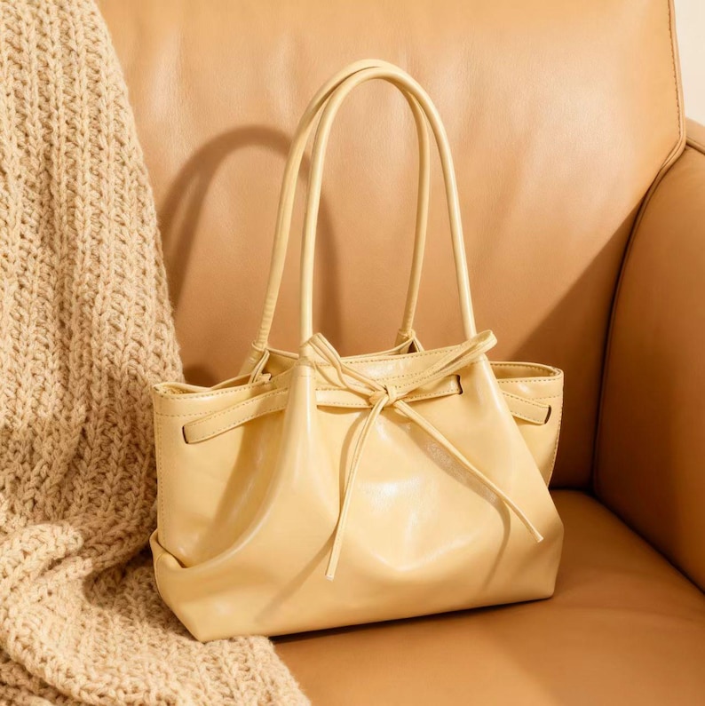 May include: A pale yellow handbag with two curved handles and a drawstring closure. The bag is made of a smooth material and has a rectangular shape with rounded corners. The bag is sitting on a tan leather chair.