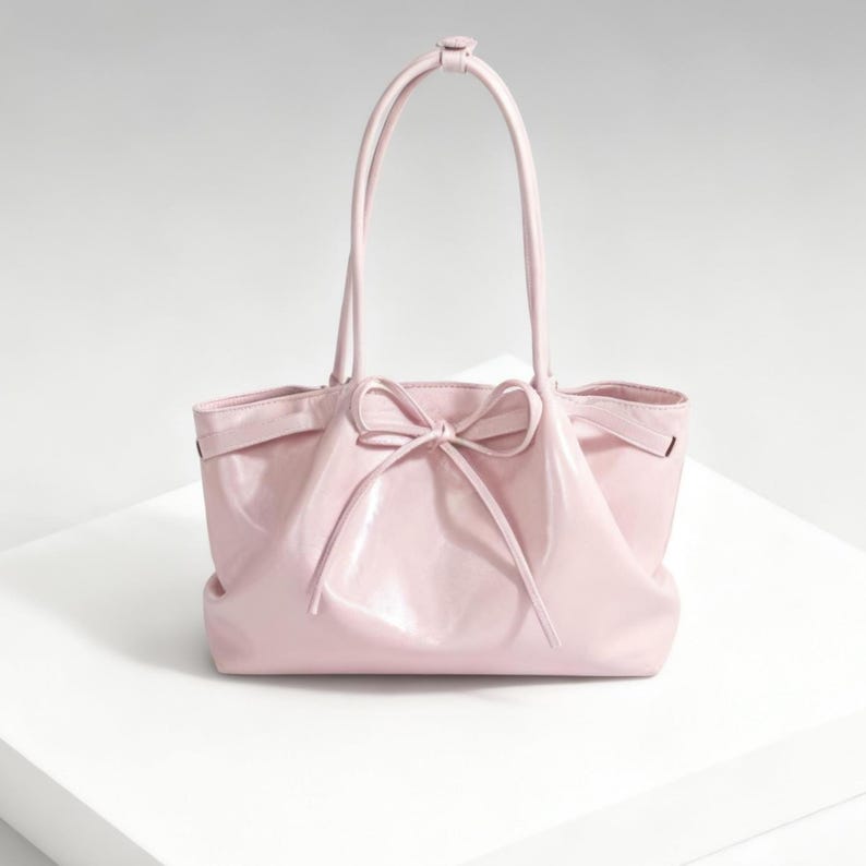 May include: A pale pink tote bag with a bow detail. The bag features two long handles and a smaller top handle. It's made from a shiny material, giving it a soft, feminine appearance.