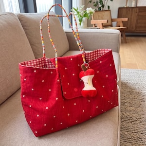 May include: A red tote bag with white floral detailing and a gingham lining. The bag has colourful beaded handles and a flap closure. An apple core charm hangs from the bag. The bag is on a beige sofa. The bag is suitable for shopping.