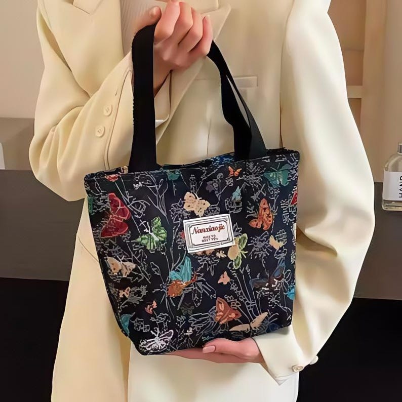 May include: A black tote bag adorned with a vibrant butterfly print. The bag has a rectangular form and short black handles. The design showcases butterflies in red, orange, green, and blue, set against a dark backdrop.