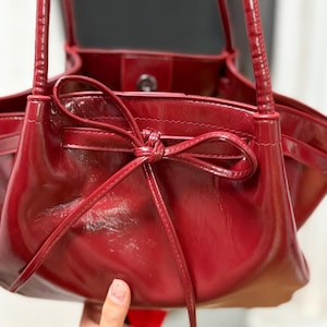 May include: A glossy, deep red handbag with a bow detail at the top. The bag has a structured shape with a smooth, shiny finish. The handles are thin and match the bag's colour. The interior is visible, and the bag appears to be made of leather or a similar material.
