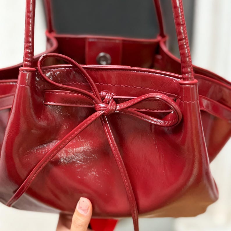 May include: A glossy, deep red handbag with a bow detail at the top. The bag has a structured shape with a smooth, shiny finish. The handles are thin and match the bag's colour. The interior is visible, and the bag appears to be made of leather or a similar material.