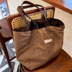 May include: A brown tote bag with a leopard print lining and handles. The bag features a front pocket with a small white label that reads "really loved". The bag is on a wooden chair.