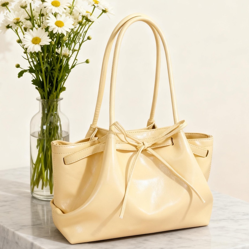 May include: A pale yellow handbag with a drawstring closure and two handles. The bag is made of a shiny material and has a simple, elegant design. A vase of white daisies sits next to the bag.