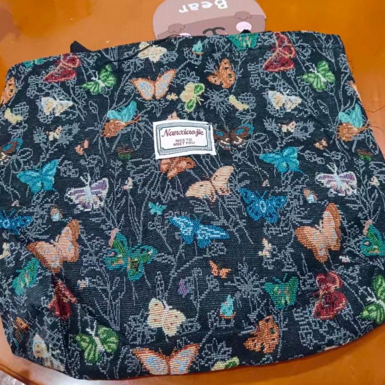 May include: A black tote bag featuring a vibrant butterfly design in shades of blue, orange, green, and red. The bag includes a label that reads "Nanciaojie, NICE TO MEET YOU".