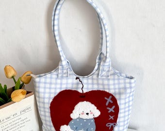 May include: A light blue and white gingham tote bag with a red heart applique featuring a white dog wearing a blue sweater. The bag has a matching gingham handle and a bone-shaped detail.