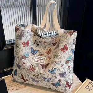 May include: Cream tote bag featuring a butterfly design in red, blue, and purple. The bag has off-white handles and a small rectangular tag with text. A stylish and practical accessory for everyday use.