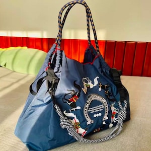 May include: A navy blue tote bag with a drawstring closure and rope handles. The bag features embroidered designs of horses and the text "Ball and Chain". The handles are a combination of navy, yellow, and white rope.