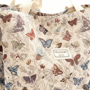 May include: Cream-coloured tote bag featuring a repeating pattern of colourful butterflies and foliage. The bag has cream-coloured handles and a small rectangular label with text. The butterflies are in shades of red, blue, brown, and purple.