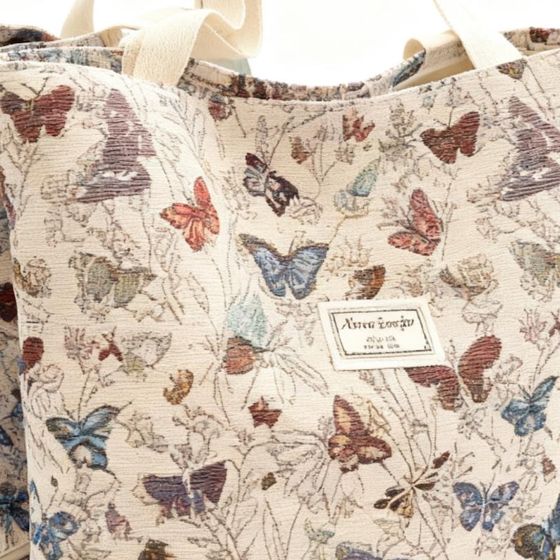 May include: Cream-coloured tote bag featuring a repeating pattern of colourful butterflies and foliage. The bag has cream-coloured handles and a small rectangular label with text. The butterflies are in shades of red, blue, brown, and purple.