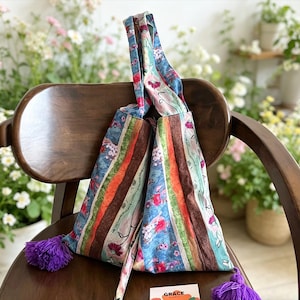 May include: A patterned tote bag in shades of blue, orange, and brown, with a floral and striped design. The bag has purple tassels and is placed on a wooden chair. The bag's design includes floral and striped patterns.