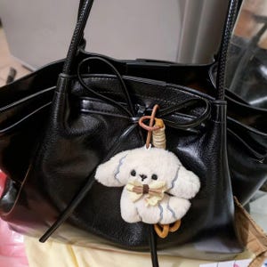 May include: A black leather tote bag with a fluffy white dog charm. The bag features long handles and a bow detail. The dog charm has a bow tie and blue detailing.