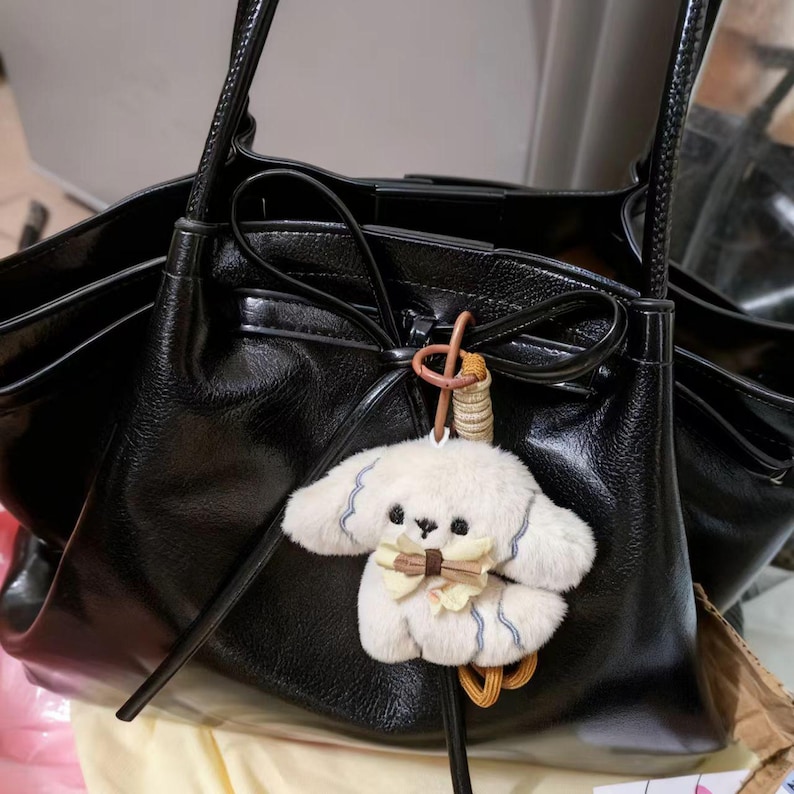 May include: A black leather tote bag with a fluffy white dog charm. The bag features long handles and a bow detail. The dog charm has a bow tie and blue detailing.