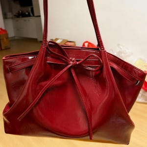 May include: A shiny, deep red tote bag with a bow detail. The bag has long handles and a spacious interior. The material appears to be patent leather or a similar shiny fabric, reflecting light. A stylish handbag.