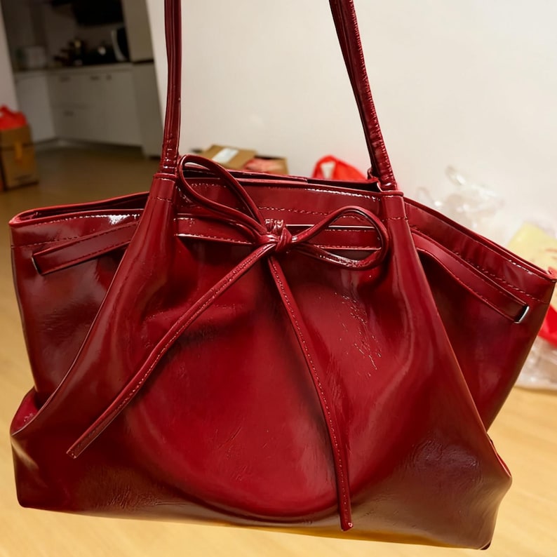 May include: A shiny, deep red tote bag with a bow detail. The bag has long handles and a spacious interior. The material appears to be patent leather or a similar shiny fabric, reflecting light. A stylish handbag.