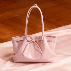 May include: A pale pink handbag featuring a bow detail. The bag has two handles and a rectangular shape. The bag is made of a glossy material and rests on a pink satin surface.