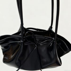 May include: A black, glossy handbag with a distinctive, gathered design. The bag has long, slender straps and a drawstring closure. The interior is visible, and the bag appears to be made of a patent leather-like material.