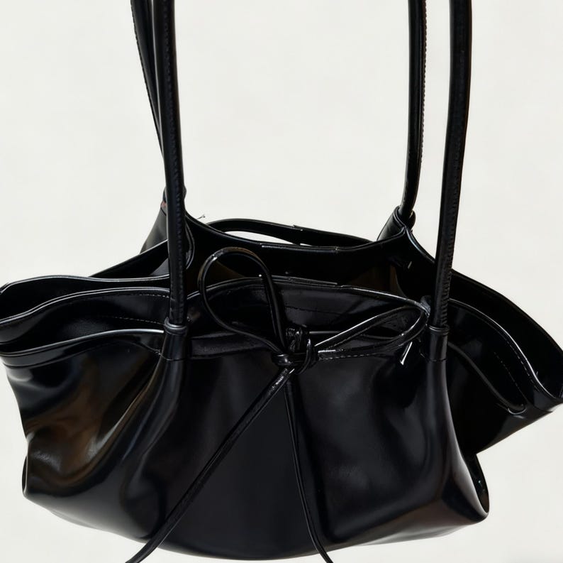 May include: A black, glossy handbag with a distinctive, gathered design. The bag has long, slender straps and a drawstring closure. The interior is visible, and the bag appears to be made of a patent leather-like material.
