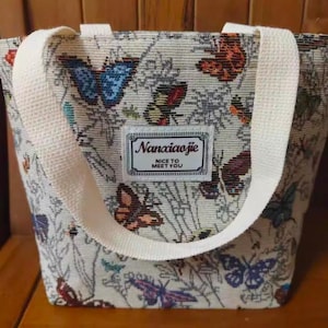 May include: Cream-coloured tote bag featuring a woven handle and a body adorned with colourful butterflies and foliage. A rectangular label on the front reads "Nanaiaojie NICE TO MEET YOU".