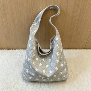 May include: A grey shoulder bag featuring a white star design. The bag has a broad strap and a button fastening. The interior lining is a pale pink hue. The bag is placed on a white, fluffy surface.