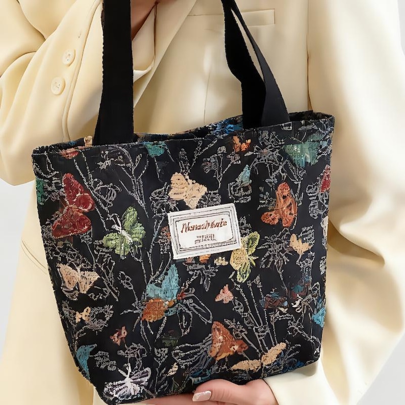 May include: Black tote bag featuring a vibrant butterfly design. The bag has black handles and a white label with text. The butterflies are in shades of red, orange, blue, green, and yellow.