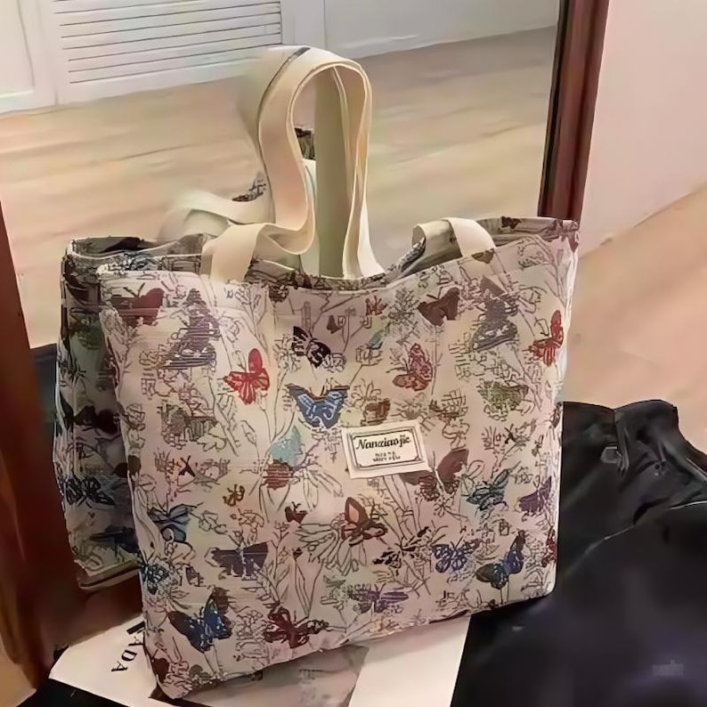 May include: A cream tote bag featuring a pattern of colourful butterflies and floral designs. The bag has cream handles and a small rectangular label with the text "Nanxiao jie".