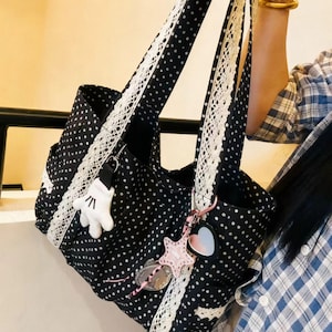 May include: Black tote bag with white polka dots, white lace trim, and matching handles. The bag features a white glove charm and a pink star-shaped keychain with a heart-shaped charm. The bag is held up by a person.